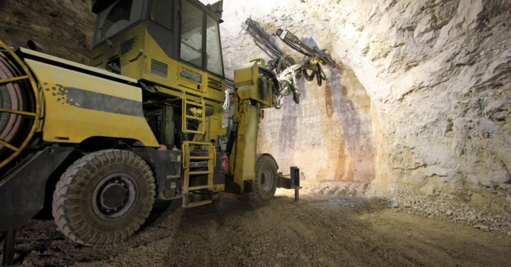 Heavy-duty equipment is in an underground tunnel of a mine. It's drilling at a rock wall as light illuminates the area.