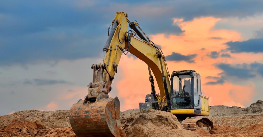 An excavator is scooping up dirt in an open-pit jobsite. The sky is cloudy with orange and blue colors.