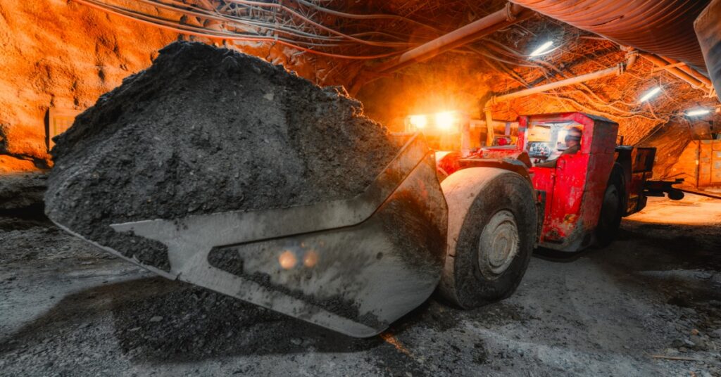 A load-haul-dump machine has a full bucket of rock and dirt in an underground mine. Orange lights illuminate the tunnel.