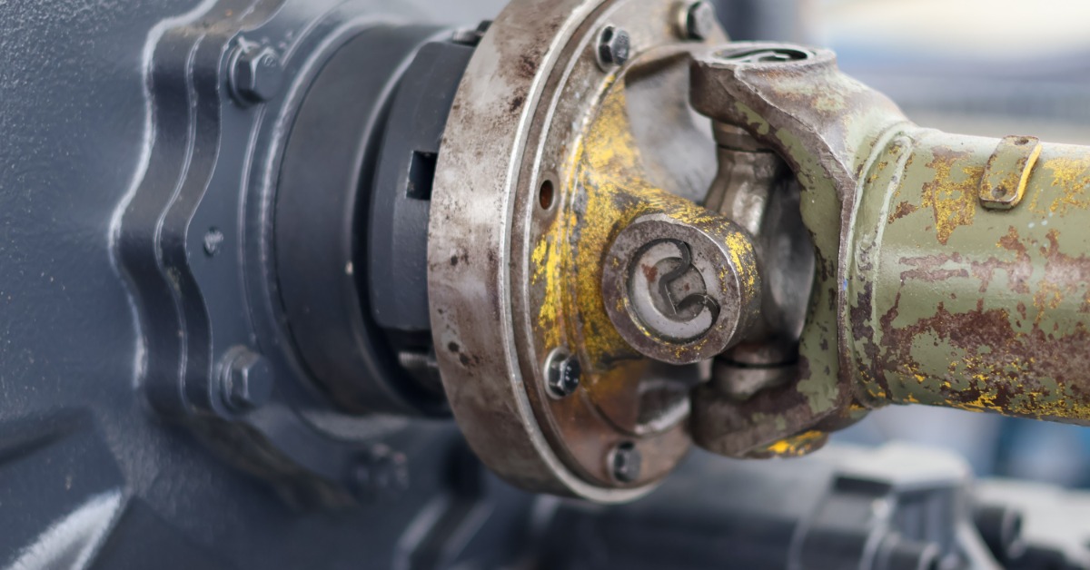 A close-up of a worn, rusted, greasy universal joint and driveshaft coupling on a piece of heavy machinery.