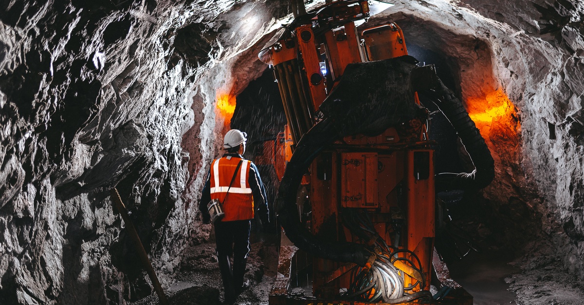 A person wearing an orange reflective vest is walking beside a drilling machine in the tunnel of an underground mine.