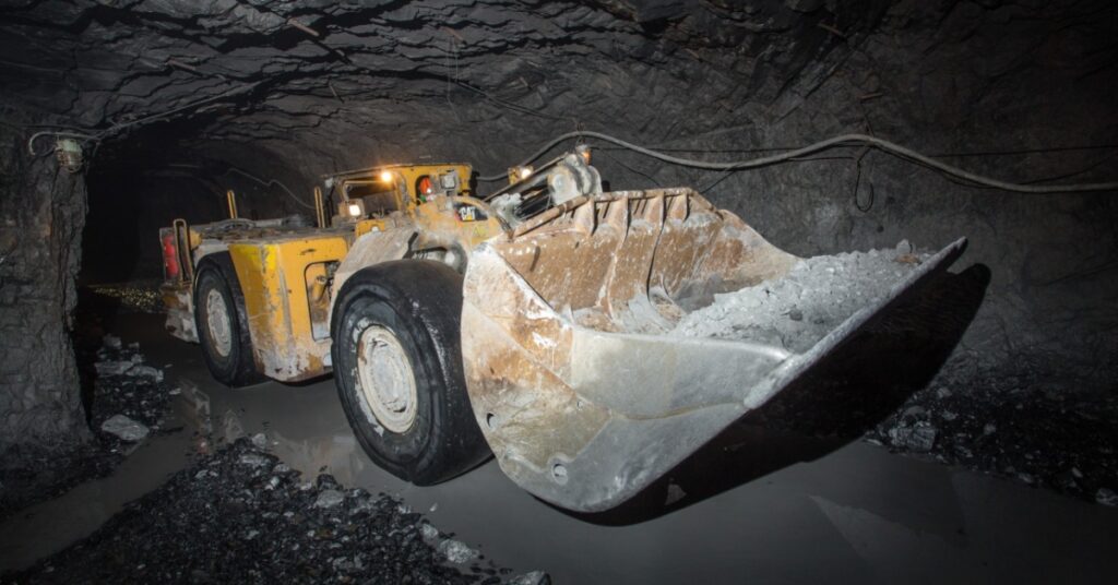 A load-haul-dump machine is carrying rock in a tunnel of an underground mine. Water and rock coat the surface.