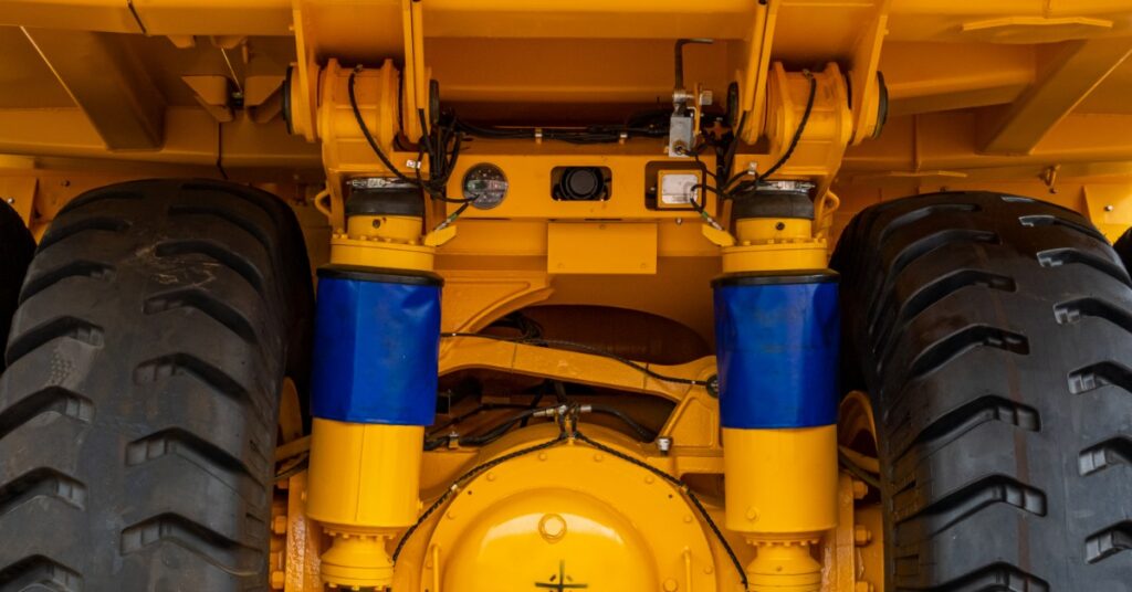A close-up view of yellow mining truck tires, axle, suspension parts, and blue hydraulic covers outdoors.