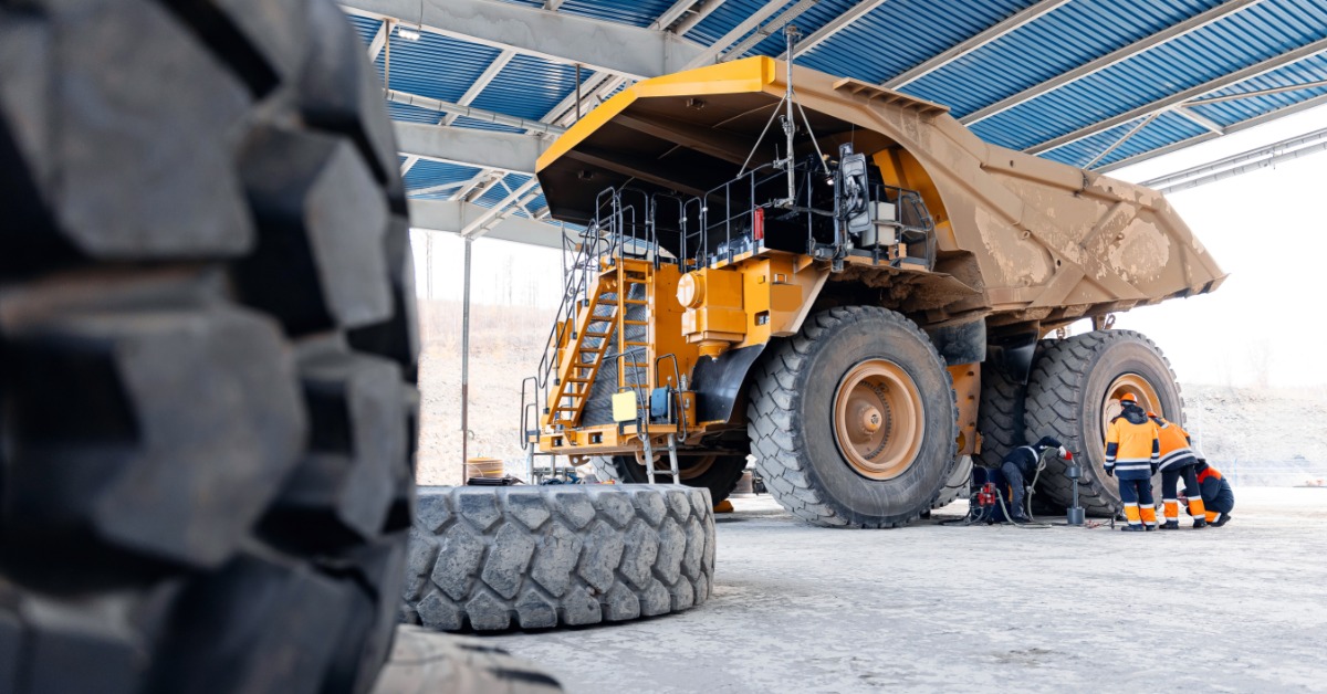 A person working near large mining truck tires inside a garage with tools and heavy vehicle parts nearby.