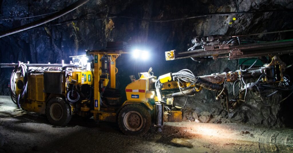 A large drilling machine is parked inside an underground mine tunnel with rocky walls and dim industrial lighting.