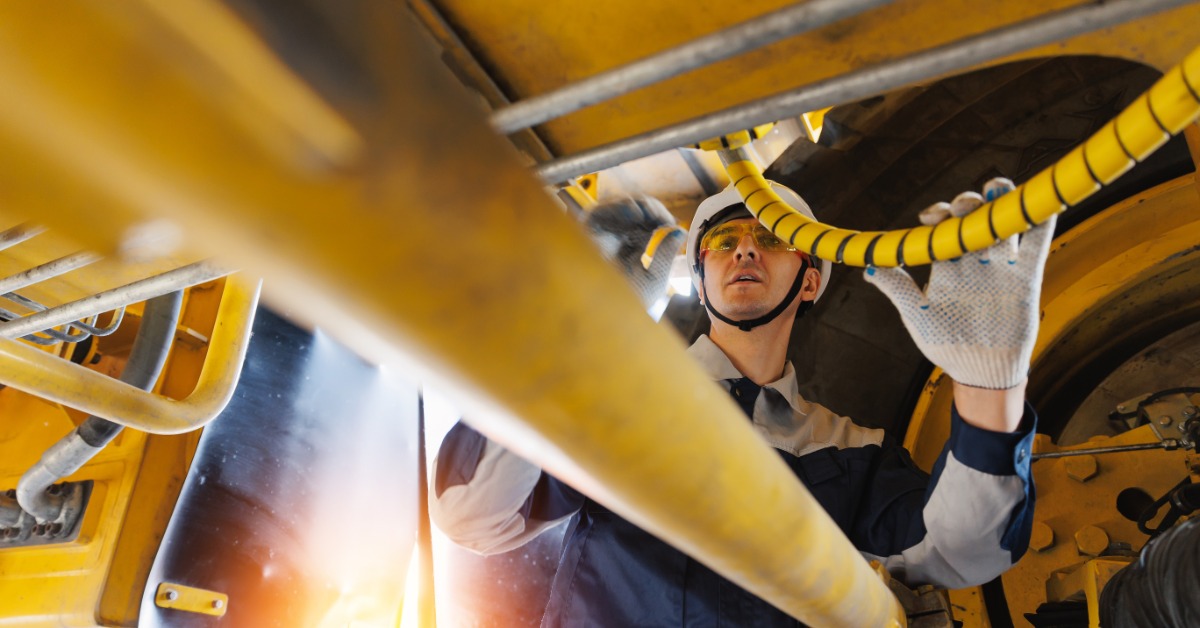 A ground-level view of a man wearing a hard hat and gloves inspecting heavy machinery. The metal components are yellow.