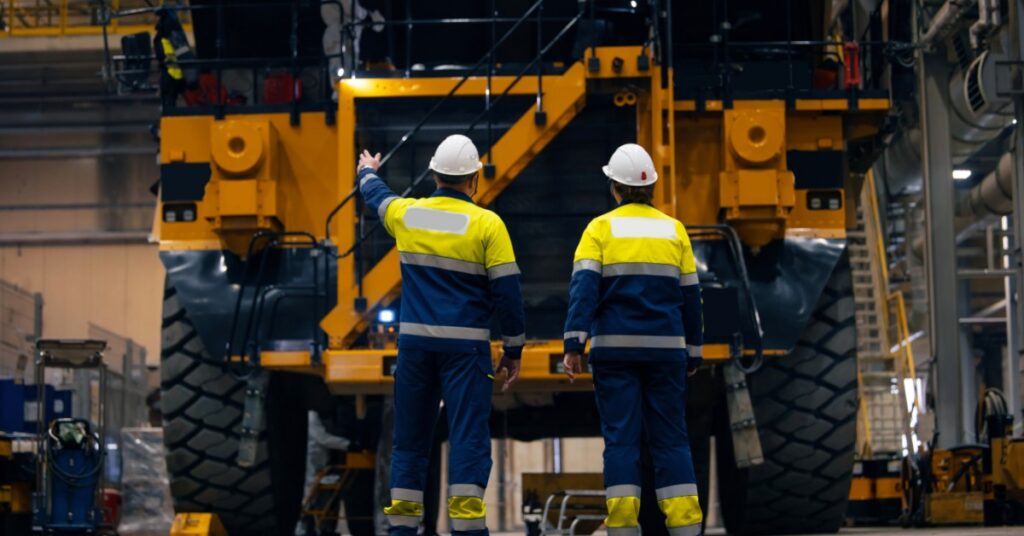 Two workers in hard hats and high-visibility uniforms are standing in front of a large yellow dump truck indoors.