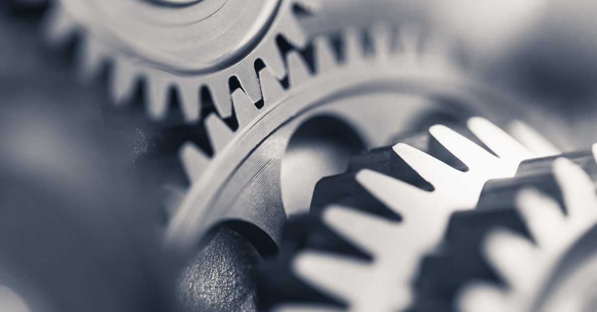 A close-up of interlocking silver metal gears with sharp teeth. Blurred metal mechanical components are in the background.