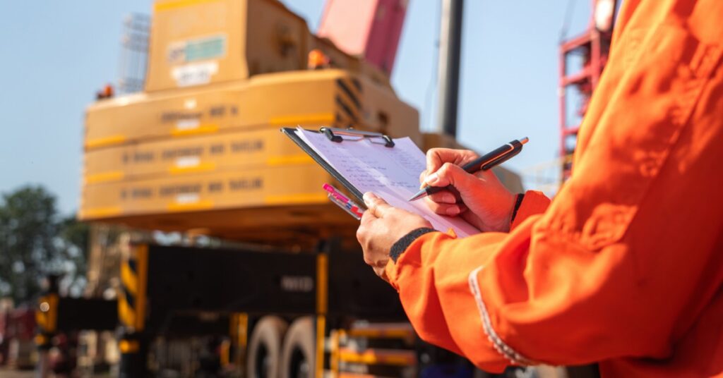 A person wearing gloves holds a clipboard while inspecting a machine. An industrial crane is blurred in the background.