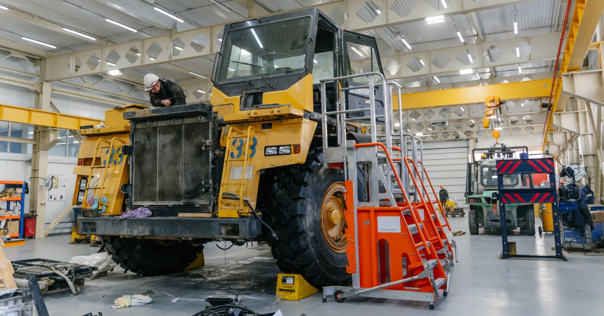 A person wearing a hard hat repairs a yellow dump truck inside a large industrial garage. Various tools lie on the floor.