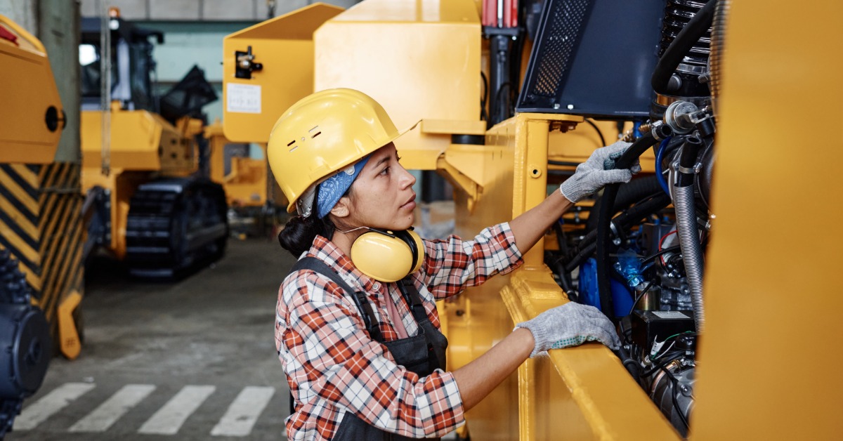 A woman in a hard hat, gloves, and coveralls checks the open engine area of a large, yellow industrial machine.