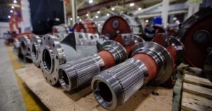 A row of large red and silver axles and spare metal parts resting on pallets inside an industrial workshop.