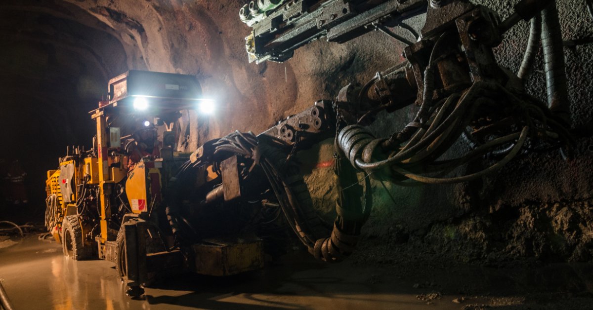 A yellow mining machine with bright headlights working beside a rock wall in a dark underground tunnel.