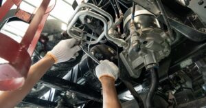 Gloved hands reaching under a truck chassis near hoses, pipes, tanks, and metal undercarriage components.