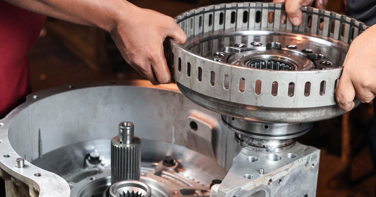 Several hands lifting a large circular metal gear hub above an open industrial transmission housing.