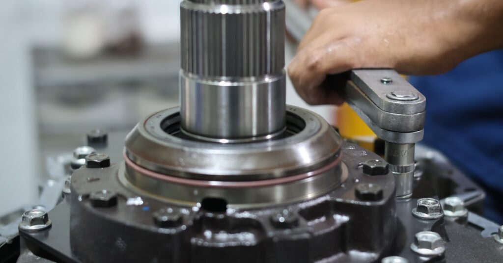 A hand using a torque wrench to tighten bolts on a metal transmission component in a workshop with visible machined holes.