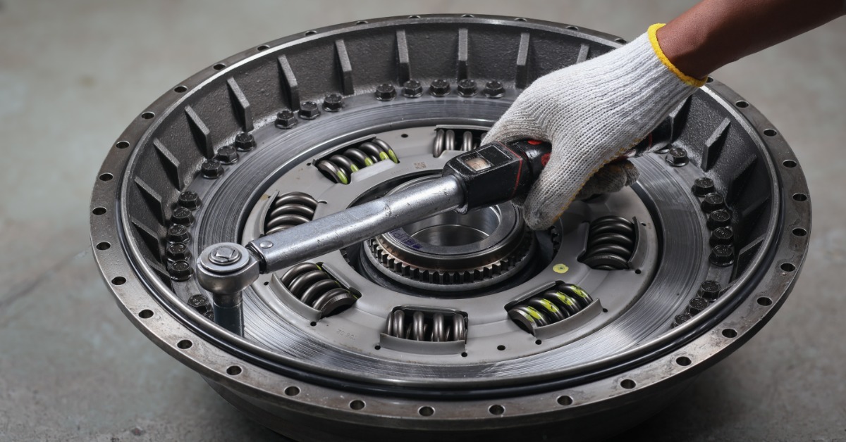 A gloved hand using a torque wrench on a circular torque converter with bolts and visible springs in a workshop.