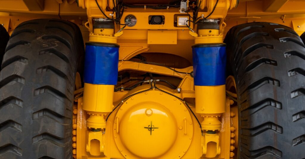 A yellow mining truck undercarriage with two black tires, blue shock covers, hydraulic lines, and a central axle housing.