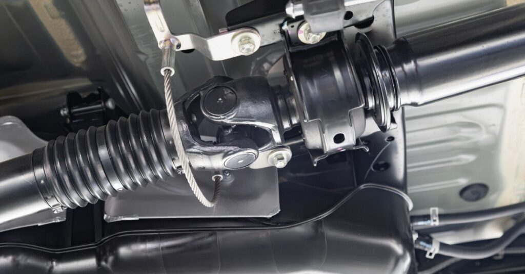 A black driveshaft with a universal joint, metal safety cable, bolts, and undercarriage parts are beneath a vehicle.