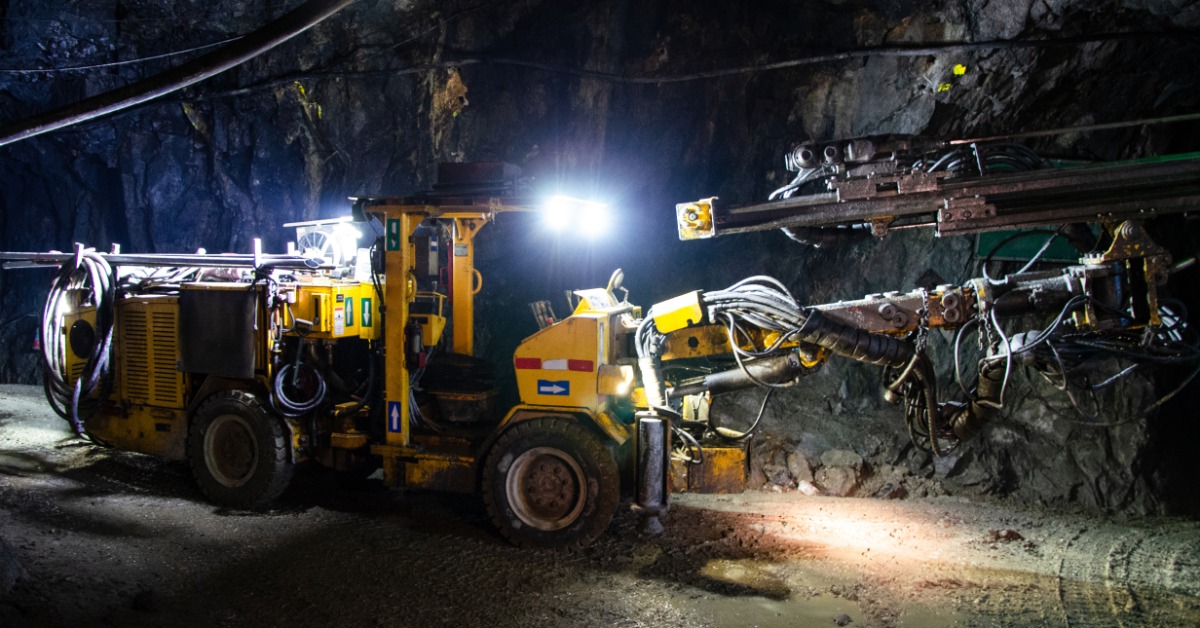 A red jumbo drill with extended booms stands inside a dark underground mine tunnel against a rough rock wall.