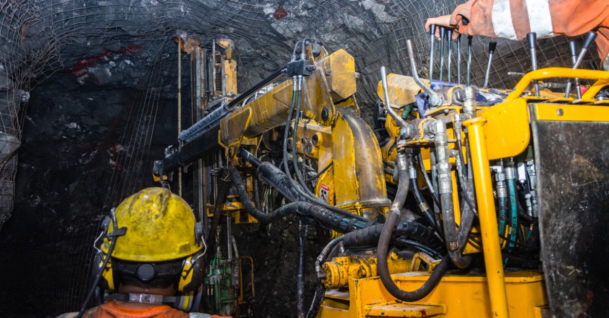 An equipment operator reaches toward controls on a mining machine in a rock tunnel. Another worker stands behind the machine.