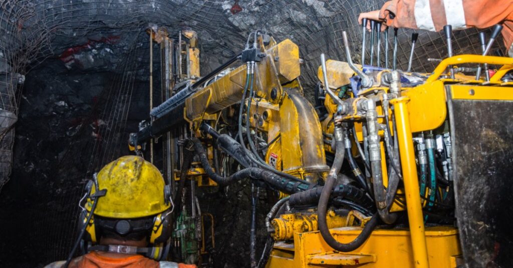 A person in orange safety gear operates yellow mining machinery beneath wire mesh in an underground rock tunnel.