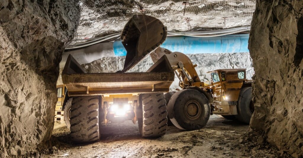 A yellow loader tipping material into a large mining truck beneath rock walls and blue ducting in an underground mine.