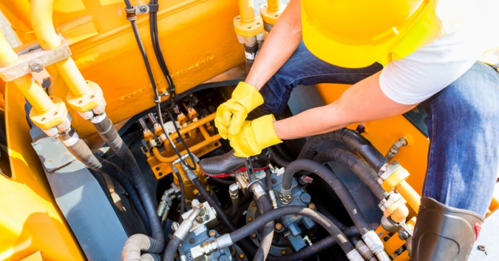 A person wearing a yellow hard hat and gloves repairing hoses on bright yellow construction machinery.