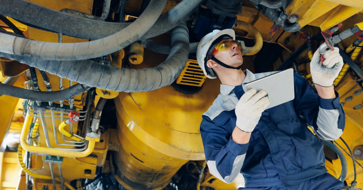 A man wearing a white hard hat and safety glasses using a tablet beside red industrial machinery outdoors.