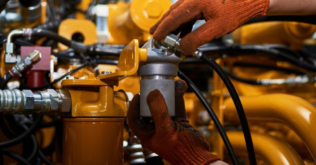 A person wearing orange work gloves tightens a metal hydraulic connector on yellow machinery with black hoses.