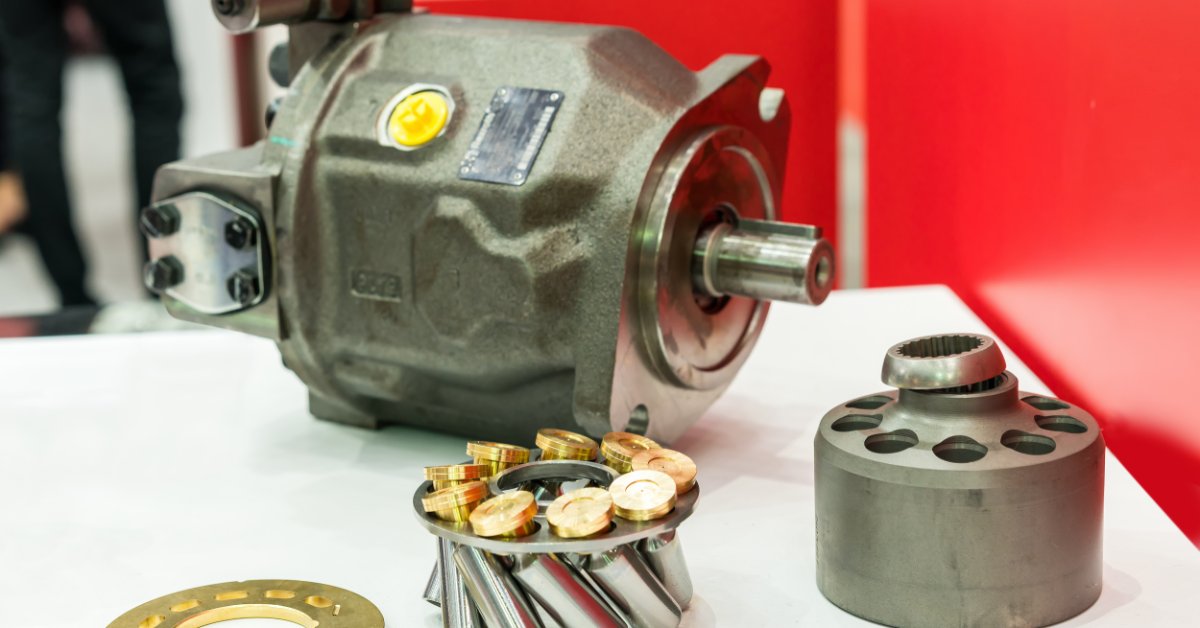 A close-up of a hydraulic pump, round cylinder, and a brass plate sitting on a white table beside metal piston parts.