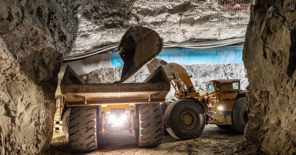 A loader pours ore into a mining truck inside a rocky tunnel with mesh, blue ducting, large tires, and headlights.