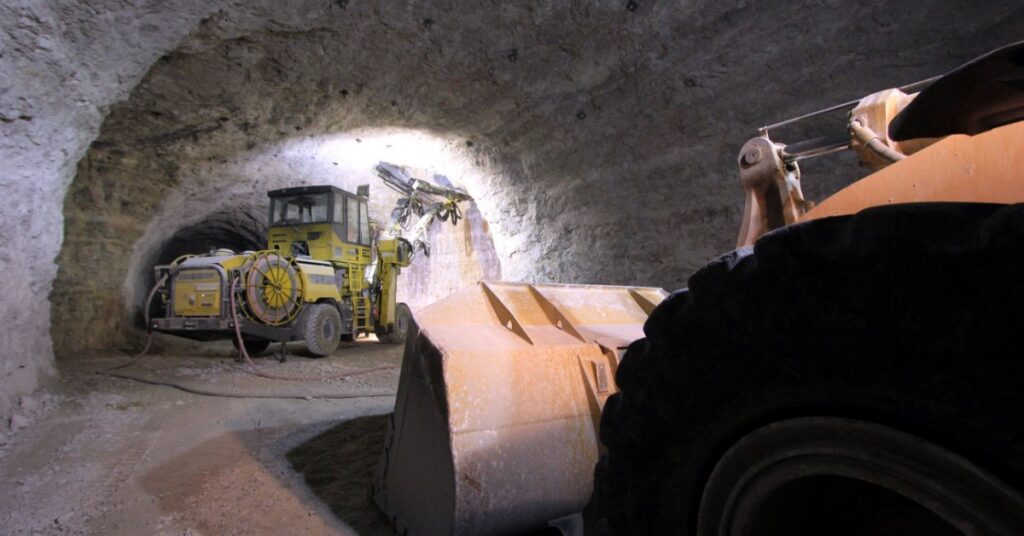 A large loader bucket and yellow drilling machine are parked inside an underground mine tunnel with rough walls.