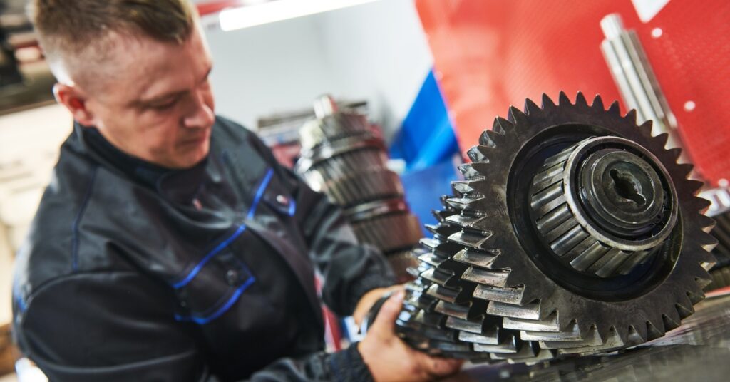 A man in a dark work jacket handling a large toothed gearbox shaft on a bench inside a brightly lit repair shop.