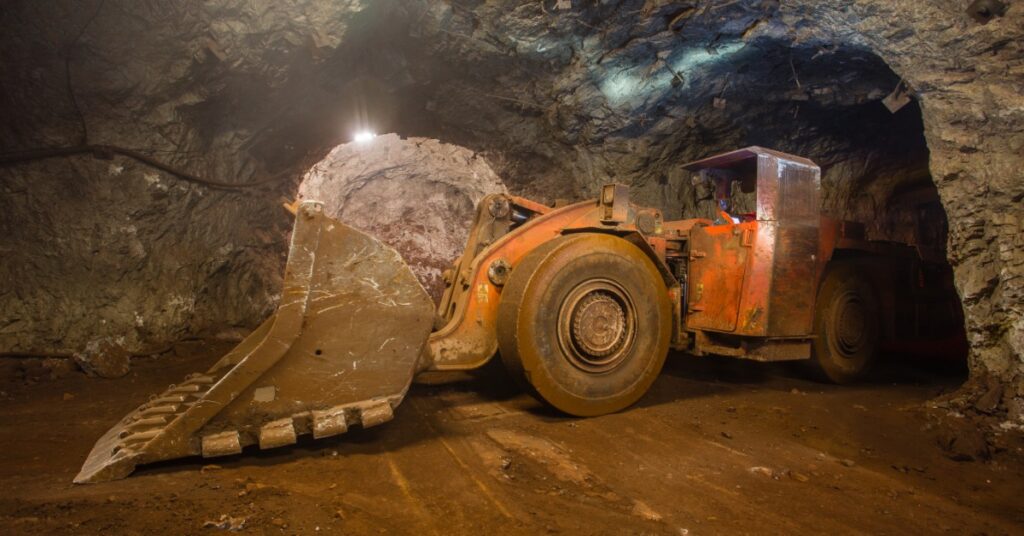 A large orange loading machine parked in a rocky underground mine tunnel with its scoop resting on the dirt floor.