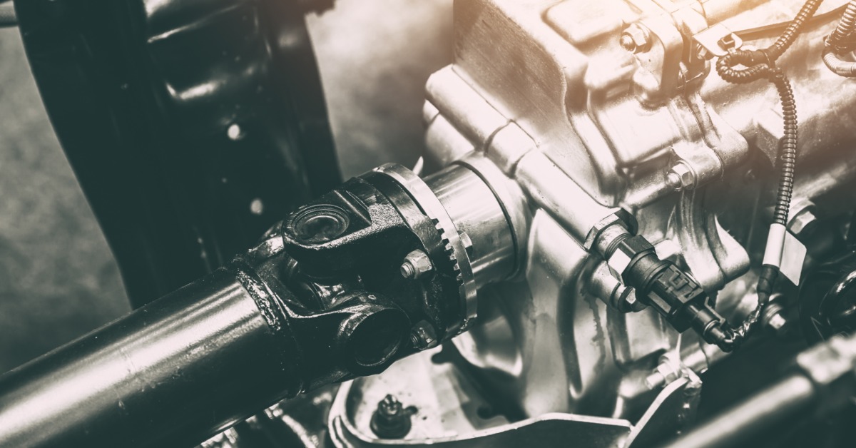 A black driveshaft and universal joint connect to a metal gearbox inside the undercarriage of heavy equipment.