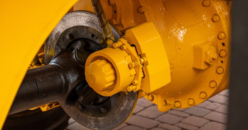 A yellow industrial axle and brake assembly shows a black driveshaft, silver disc, bolts, and hydraulic line.